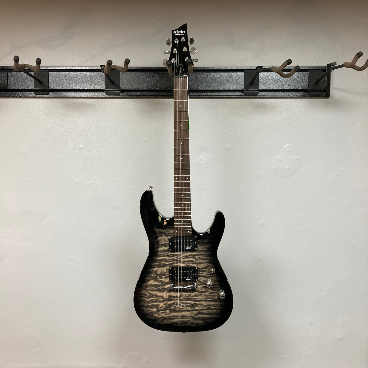 Schecter C-6 Plus Charcoal Burst Electric Guitar with green pickguard, displayed on a wall hook. Features bolt-on maple neck and Schecter open-coil humbuckers.